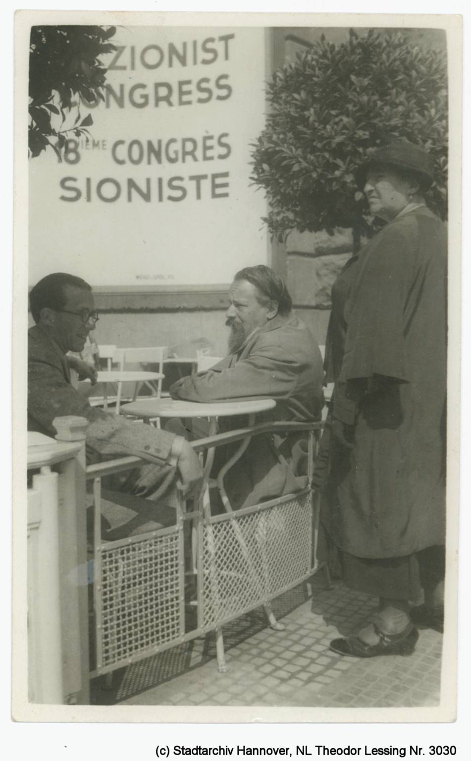 Mann sitzt mit einem anderen Mann draußen an einem Café-Tisch, daneben steht eine Frau. Im Hintergrund ist ein Schild mit der Aufschrift "Zionisten-Kongress" zu sehen.
